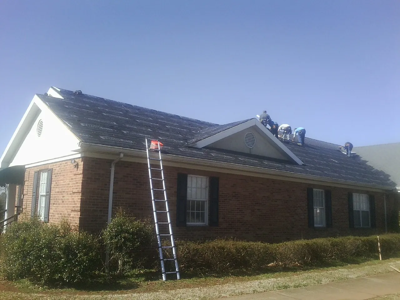 Crew tearing shingles off a brick ranch roof with a ladder set — Emergency Roof Repair work by our crew in Cairnbrook