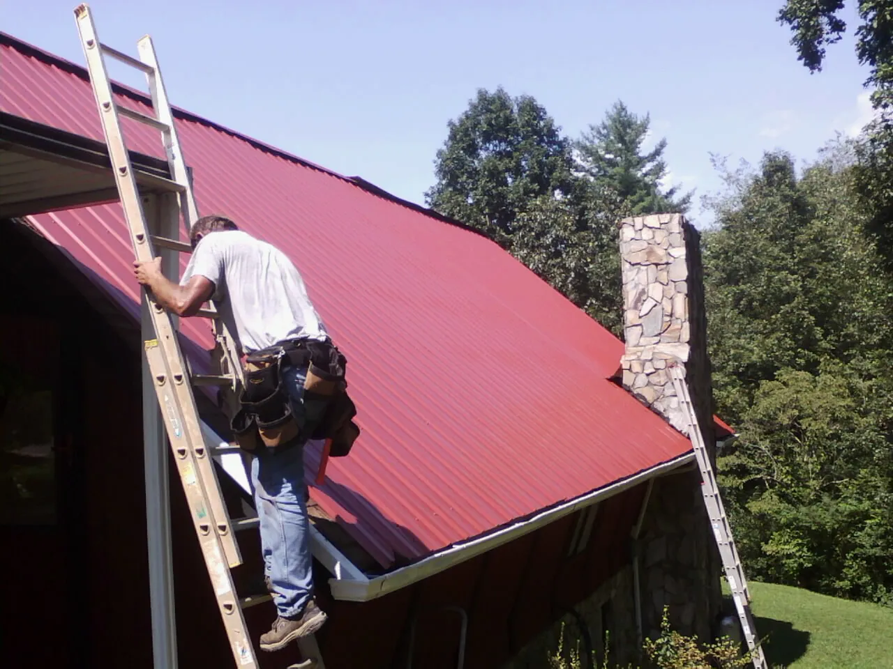 Roofer on a ladder beside a red metal roof and stone chimney — Soffit & Fascia Repair by Roofing Brothers in Cairnbrook